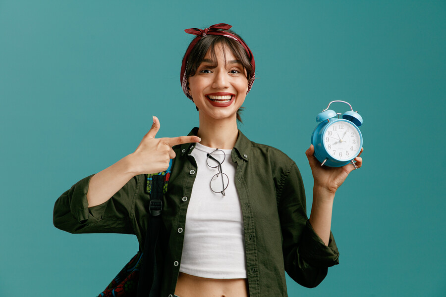joyful-young-student-girl-wearing-bandana-backpack-looking-camera-showing-alarm-clock-pointing-it-isolated-blue-background_2882c.jpg