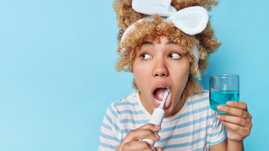 studio-shot-surprised-curly-haired-young-woman-looks-away-brushes-teeth-takes-care-oral-hygiene-uses-electric-brush-holds-mouthwash-poses-against-blue-background-blank-space-your-text_32ca4.jpg
