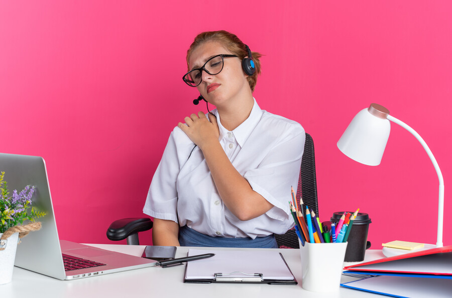 aching-young-blonde-call-centre-girl-wearing-headset-glasses-sitting-desk-with-work-tools-looking-laptop-keeping-hand-shoulder-isolated-pink-wall_2ee8f.jpg
